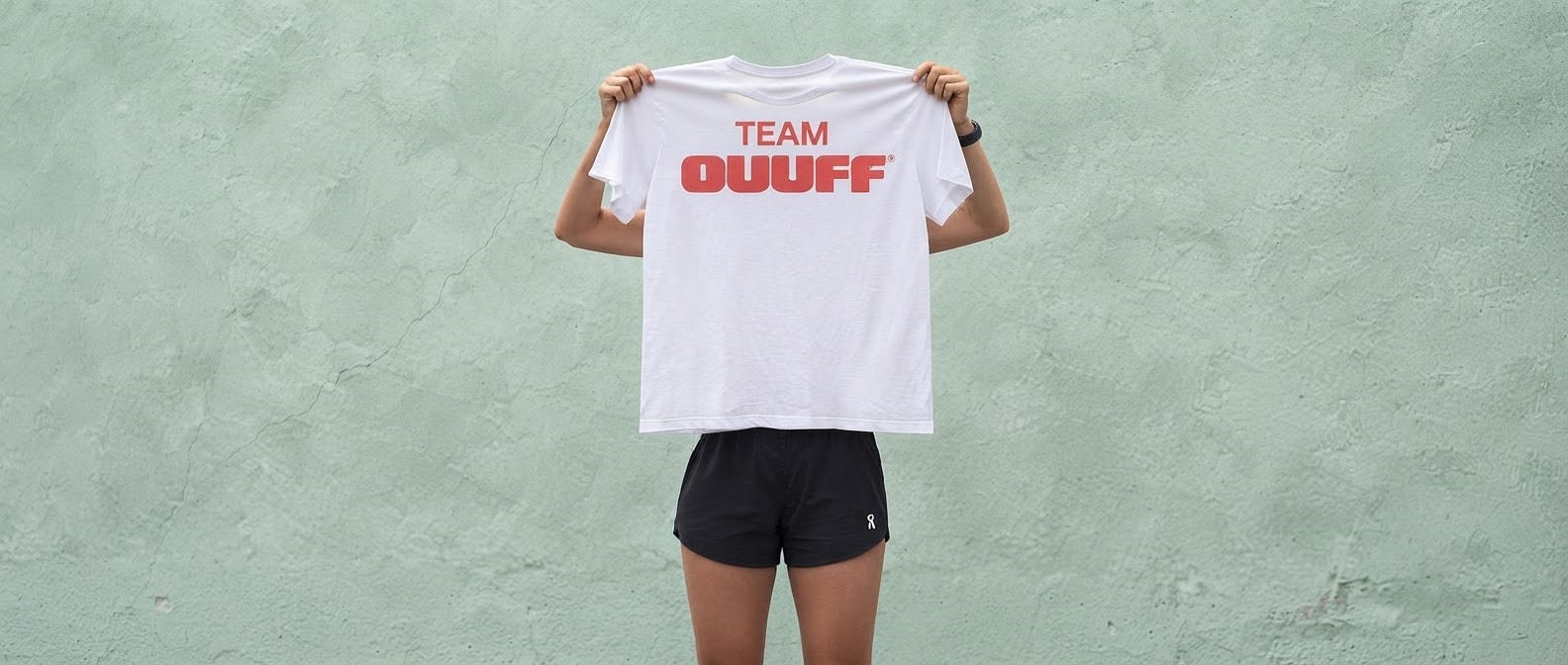 Someone holding up a white TEAM OUUFF T-shirt in front of a mint green wall